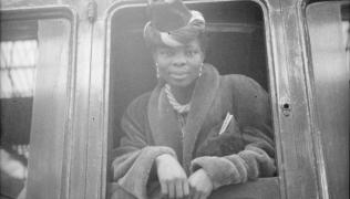 A black and white photograph of Princess Omo-Oba Adenrele Ademola looking out the door of a train. She wears a hat and fur coat.
