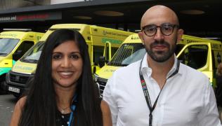 Dr Anjali Chander and Dr Miguel Reis Ferreira stood next to each other, smiling, outside Guy’s Hospital. Behind them are some ambulances in front of the hospital entrance. .