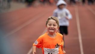 A young patient running on a race track. They are wearing an orange t-shirt that has an 'Evelina VIPs' logo in the top right corner, and and 'Evelina London Children's Charity' logo on the left sleeve. They are wearing competitor number 56 and smiling. 