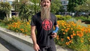 Marcin is wearing a black t-shirt with a red logo on the front, a red cap and black trousers. He is standing in St Thomas' hospital gardens, with plants behind him. He has a blue bandage on his left hand.