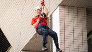 Nigel Haselden abseiling down St Thomas' Hospital. He is wearing a red Guy's & St Thomas' Charity t-shirt and a white helmet. 