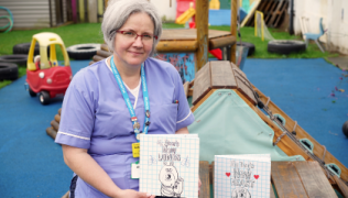 Image of Jola holding a transplant activity book. She is sitting in an outdoor area of a nursery. Jola is wearing a nurse's uniform, glasses and a blue lanyard. Next to her is another transplant activity book.