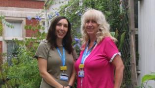 Two nurses, are standing next to each other in a garden with Wisteria in the background. Paula is wearing a beige dress with short sleeves, Phillipa has a pink short sleeved top on.