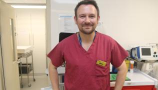 A man in claret soloured scrubs is standing in front of equipment in theatres. He wears a yellow badge marked'Alex'