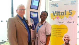 Two people stand in front of a kiosk. Next to them is a stand marked 'Vital 5