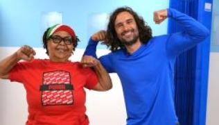 Patient is wearing a red t-shirt and cap. She has her hands in fists, up at her shoulders. Joe Wicks is standing next to her, in a blue top with his fists also up to show strength