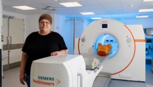 The patient is wearing a black t-shirt and brown head wrap. She is standing next to a white scanning machine, which is branded Siemens Healthineers