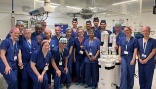 A team of surgeons wearing blue scrubs are standing in a surgical room, smiling at the camera. They are stood next to the Versius surgical robot.