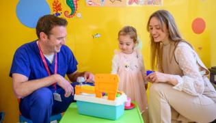 A young transplant patient sits at a table with toys in Evelina London Children's Hospital. Her mother and a doctor are sitting next to her and smiling.