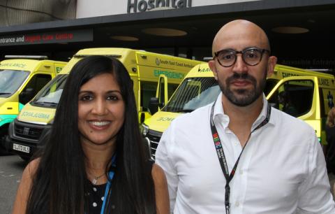 Dr Anjali Chander and Dr Miguel Reis Ferreira stood next to each other, smiling, outside Guy’s Hospital. Behind them are some ambulances in front of the hospital entrance. .