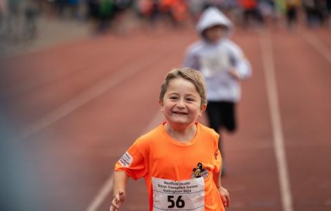 A young patient running on a race track. They are wearing an orange t-shirt that has an 'Evelina VIPs' logo in the top right corner, and 'Evelina London Children's Charity' logo on the left sleeve. They are wearing competitor number 56 and smiling.