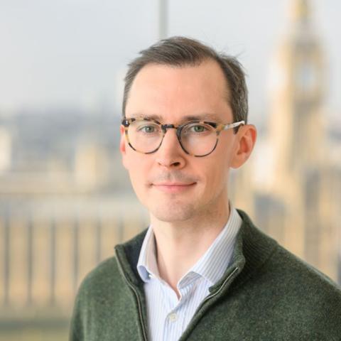 Headshot of consultant plastic surgeon, Mr George Murphy. He is wearing a pale blue shirt, with a green jumper on top - the zip of which is open at his neck. He has tortoiseshell glasses and brown hair.