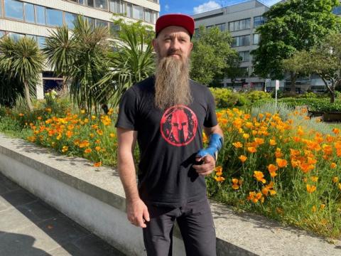 Marcin is wearing a black t-shirt with a red logo on the front, a red cap and black trousers. He is standing in St Thomas' hospital gardens, with plants behind him. He has a blue bandage on his left hand.