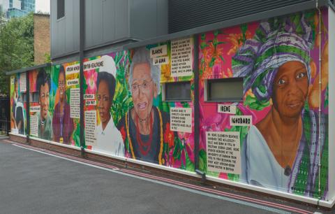 A photo of the mural celebrating African women in healthcare at Guy's Hospital. The colourful artwork, which has a background of medicinal plants is on the side of a building by a street. It features six women - Matilda Clerk, Dzagbele Matilda Asante, Metian Parsanka, Kofoworola Abeni ‘Ivy’ Pratt, Blanche La Guma, and Dr Irene Elizabeth Beatrice Ighodaro.