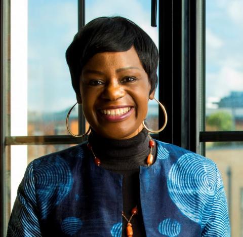 Headshot of Dr Dela Idowu, wearing a blue top, a necklance and large hoop earrings.