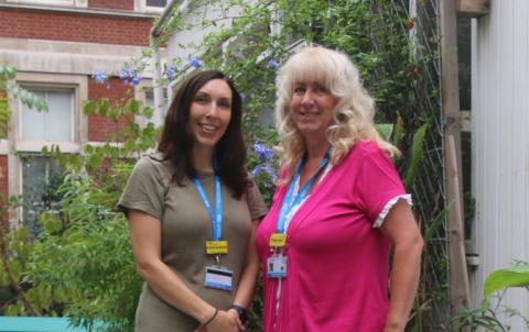 Two nurses, are standing next to each other in a garden with Wisteria in the background. Paula is wearing a beige dress with short sleeves, Phillipa has a pink short sleeved top on.