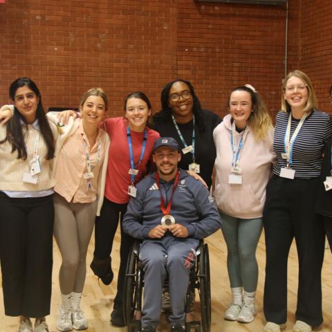 Paralympian and tennis champion, Andy Lapthorne, sitting in a wheelchair with a medal around his chest. He is surrounded by a group of 8 individuals.