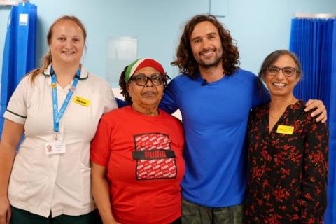 OT Abbie is wearing a white work tunic, patient has a red t-shirt on, Joe is wearing a blue top and Jugdeep a red and black dress. They are standing next to one other and smiling