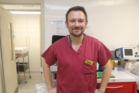 A man in claret soloured scrubs is standing in front of equipment in theatres. He wears a yellow badge marked'Alex'