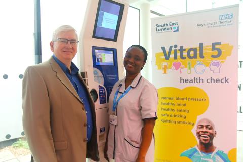 Two people stand in front of a kiosk. Next to them is a stand marked 'Vital 5