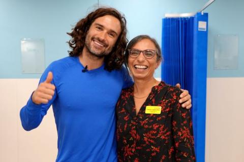 Joe Wicks is wearing a blue top. He has one thumb up and his arm around the shoulders of Prof Jugdeep Dhesi, who has a black and red dress on and is smiling.