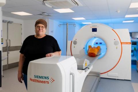 The patient is wearing a black t-shirt and brown head wrap. She is standing next to a white scanning machine, which is branded Siemens Healthineers