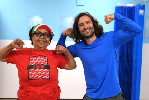 Patient is wearing a red t-shirt and cap. She has her hands in fists, up at her shoulders. Joe Wicks is standing next to her, in a blue top with his fists also up to show strength