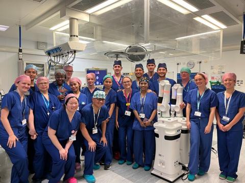 A large team of surgeons in blue scrubs are standing together in a surgical room and smiling at the camera. They are stood next to the Versius surgical robot.