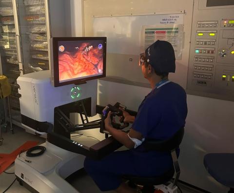 A surgeon is operating a control panel to control the Versius surgical robot. He is a dimly lit surgical room and he is looking at a screen which displays a camera image inside a patient.