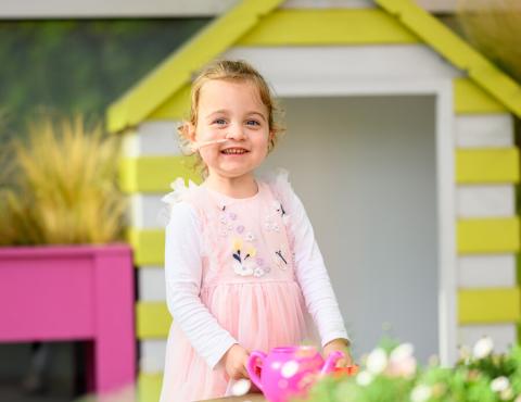 A young transplant patient in a pink dress is standing in a garden space outside Evelina London Children's Hospital. She is holding a toy teapot and smiling at the camera.