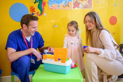 A young transplant patient sits at a table with toys in Evelina London Children's Hospital. Her mother and a doctor are sitting next to her and smiling.