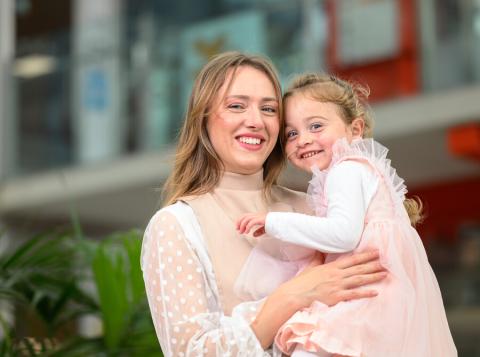 A young transplant patient in a pink dress is being held in her mother's arms inside Evelina London Children's Hospital. They are both looking at the camera and smiling.