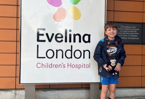 A young patient stands outside of the Evelina London hospital sign. He is in a football kit and is smiling at the camera.